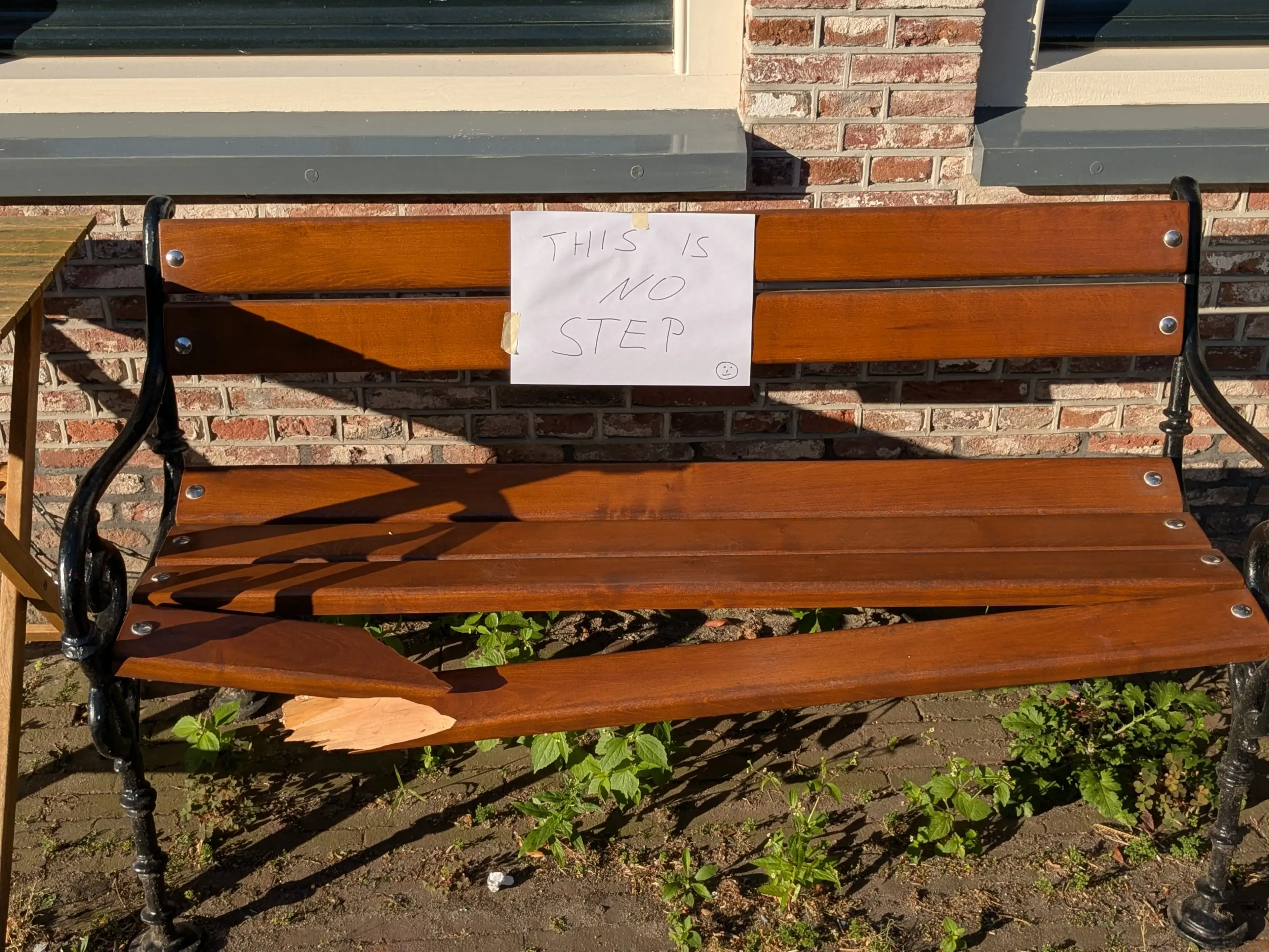 A wooden bench with one of its planks cracked in two. A piece of paper is taped to the bench which states: THIS IS NO STEP :)