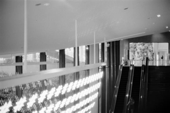 Fujifilm NEOPAN F (FF)

English
A black-and-white interior shot of a modern building with a high ceiling and large windows. The windows allow natural light to stream in, creating sharp contrasts and reflections on the glass surfaces. An escalator is visible on the right side, leading upwards. The walls are smooth and minimalist, with a large abstract art piece or sculpture visible in the background.
中文
一张黑白室内照片，展现了一个现代建筑的内部，天花板高且窗户巨大。窗户让自然光线流入，在玻璃表面形成强烈的对比和反射。右侧可见一个向上的自动扶梯。墙壁光滑且简约，背景中有一件大型抽象艺术品…