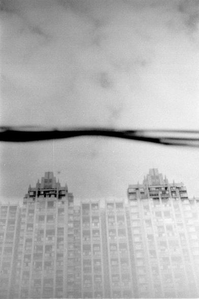 Fujifilm NEOPAN F (FF)

English
A black-and-white photograph taken from a low angle, capturing the tops of two tall buildings. The buildings have a grid-like pattern of windows, and the sky above is overcast, creating a moody atmosphere. A dark, curved object, possibly a bridge or structure, spans horizontally across the upper part of the image.
中文
一张从低角度拍摄的黑白照片，捕捉到两座高楼的顶部。建筑物的窗户呈网格状排列，上方的天空阴云密布，营造出一种沉静的氛围。上方横跨着一个黑色的弯曲物体，可能是一座桥或结构。
