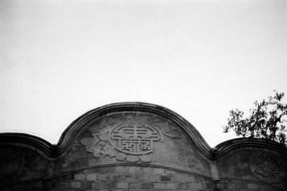 Fujifilm NEOPAN F (FF)

English
A black-and-white photograph of the upper section of a traditional Chinese architectural structure. The image focuses on a curved, decorative roof with a central carved emblem featuring the Chinese character surrounded by intricate patterns. The roof is made of gray bricks, and a tree is partially visible on the right side.
中文
一张黑白照片，拍摄的是中国传统建筑的上部。图片聚焦于一个弯曲的装饰性屋顶，中央有一个雕刻图案，周围环绕着精美的花纹。屋顶由灰色砖块构成，右侧可见部分树木。