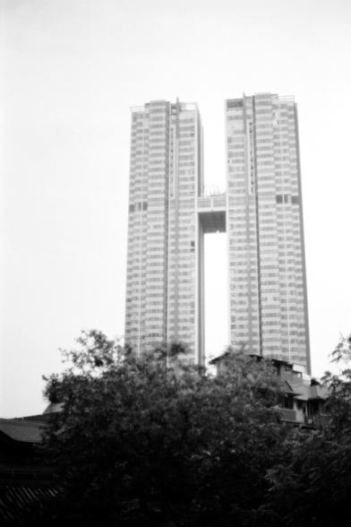 Fujifilm NEOPAN F (FF)

English
A black-and-white photograph of two tall, modern high-rise buildings connected by a horizontal bridge-like structure near the top. The buildings have numerous windows and balconies. In the foreground, there are trees and the rooftops of lower buildings, partially obscuring the base of the towers.
中文
一张黑白照片，拍摄的是两座高大的现代摩天大楼，顶部通过一个水平的桥梁状结构连接。大楼有众多窗户和阳台。前景中有树木和较低建筑的屋顶，部分遮挡了塔楼的底部。