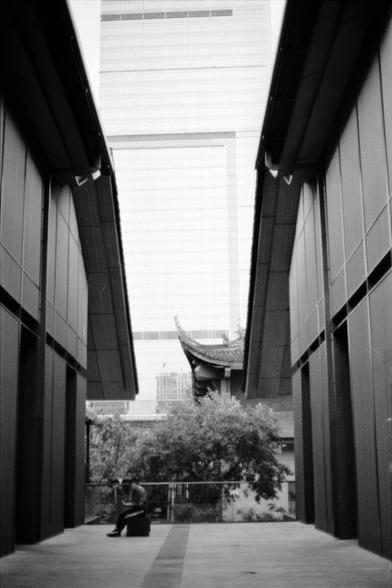 Fujifilm NEOPAN F (FF)

English
A black-and-white photograph of a narrow alleyway flanked by tall, modern buildings with smooth, dark facades. At the end of the alley, a person is seated on a bench facing away from the camera. In the background, there is a traditional Chinese building with a curved roof, surrounded by trees.
中文
一张黑白照片，拍摄的是一条狭窄的小巷，两侧是高大的现代建筑，外墙光滑且颜色深暗。小巷尽头，一个人背对着相机坐在长椅上。背景中有一座传统中式建筑，屋顶弯曲，周围环绕着树木。
