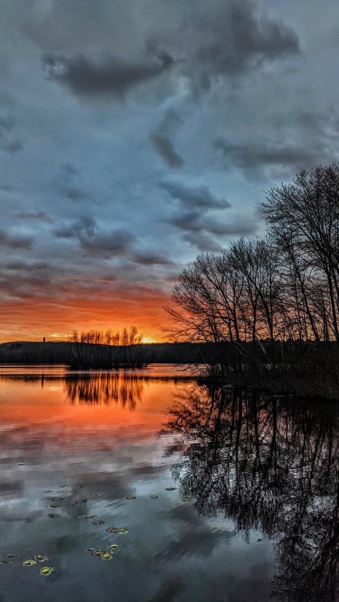 We are standing beside a large lake surrounded by a forest of leafless trees waiting for spring. The sky is overcast with thick dark gray clouds, but small breaks in the clouds along the horizon allow the rays of the recently set sun to paint the bottoms of the clouds near the horizon a vibrant orange. The sky and trees along the shore are mirrored on the lake surface.
