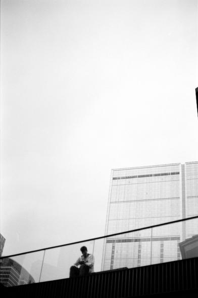 Fujifilm NEOPAN F (FF)

English
A black-and-white photograph showing a person sitting alone on a glass-railed balcony or terrace. The individual is positioned on the left side, facing slightly to the right, and appears to be looking down or at something in their hands. In the background, a tall, modern building with a grid-like facade is visible under a bright, overcast sky.
中文
一张黑白照片，展示了一个独自坐在玻璃栏杆阳台或露台上的人。该人物位于左侧，略微向右转身，似乎在低头或查看手中的物品。背景中，一幢带有网格状立面的现代高层建筑在明亮的阴天下清晰可见。