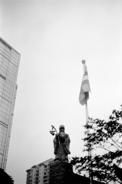 Fujifilm NEOPAN F (FF)

English
A black-and-white photograph featuring a religious statue of a robed figure holding a cane. The statue stands on a pedestal, with a flagpole to its right flying a flag. In the background, there are tall buildings, including one with a grid-like facade, and trees framing the scene.
中文
一张黑白照片，展示了一尊手持木杖身着长袍的宗教人物雕像。雕像立在基座上，右侧有一根旗杆，悬挂着一面旗帜。背景中有高层建筑，其中一幢建筑带有网格状立面，还有树木点缀着场景。