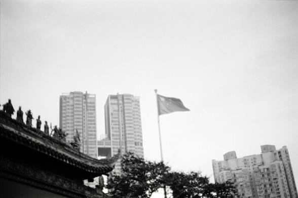 Fujifilm NEOPAN F (FF)

English
A black-and-white photograph capturing a traditional Chinese temple roof with intricate carvings and statues of figures lined up along the ridge. In the background, modern high-rise buildings are visible, and a flag with a star design is flying on a flagpole to the right of the temple roof.
中文
一张黑白照片，拍摄了一个传统中国寺庙的屋顶，屋脊上排列着精美雕刻和多尊人物雕像。背景中可见现代高层建筑，寺庙屋顶右侧的旗杆上悬挂着一面带有星形图案的旗帜。