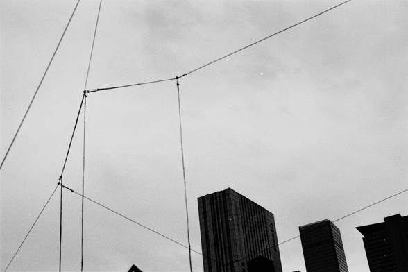 Fujifilm NEOPAN F (FF)

English
A black-and-white photograph showing a network of overhead power lines or cables against a cloudy sky. Below the lines, the tops of several tall, modern buildings are visible, creating a contrast between the industrial elements and the urban architecture.
中文
一张黑白照片，展示了在多云天空下的架空电线或电缆网络。电线下方可见多幢现代高层建筑的顶部，形成工业元素与城市建筑之间的对比。