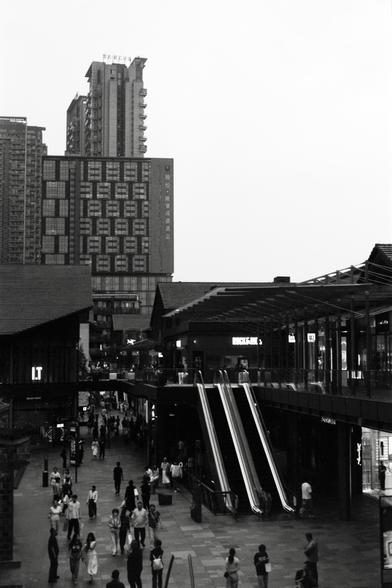 Fujifilm NEOPAN F (FF)

English
A black-and-white photograph of a bustling urban scene. The foreground features a pedestrian plaza with people walking in various directions. To the right, there are escalators leading up to a covered walkway or building entrance. In the background, tall buildings with numerous windows and signage in Chinese characters are visible. The sky is overcast, and the overall atmosphere is lively and busy.
中文
一张黑白照片，展现了繁忙的城市景象。前景是一个行人广场，人们朝不同方向行走。右侧有自动扶梯通往一个有顶棚的步行道或建筑入口。…