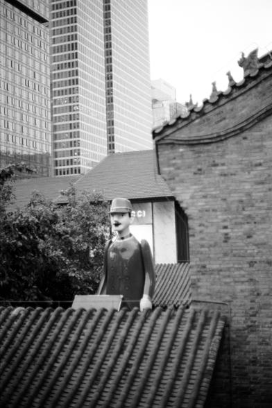 Fujifilm NEOPAN F (FF)

English
A black-and-white photograph featuring a statue of a uniformed figure, possibly a guard or official, standing on a rooftop. The statue wears a hat and a double-breasted jacket. Behind the statue, there is a traditional Chinese building with ornate roof details and a modern skyscraper in the distant background. Trees and foliage are visible to the left.
中文
一张黑白照片，展示了一个穿制服的人物雕像，可能是卫兵或官员，站在一个屋顶上。雕像戴着帽子，穿着双排扣上衣。雕像后方是一幢传统中式建筑，屋顶装饰精美，远处背景中还有一幢现代摩天大楼。左侧可见树木和绿植。