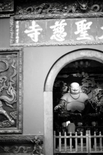 Fujifilm NEOPAN F (FF)

##English
A black-and-white image of the entrance to a temple or shrine. Above the arched doorway, there is a horizontal signboard with Chinese characters "寺" (temple) and other text. Inside the archway, a statue of a laughing Buddha is visible, surrounded by decorative elements and a fence. The walls on either side of the entrance are adorned with intricate carvings.
##中文
一张黑白照片，显示一座寺庙或神社的入口。拱形门上方有一块横向的牌匾，上面有“寺”字和其他中文字样。拱门内可见一尊笑佛雕像，周围有装饰元素和栅栏。入口两侧的墙壁上饰有精美的浮雕。