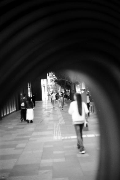Fujifilm NEOPAN F (FF)

##English
A black-and-white image of a covered walkway or tunnel with a curved ceiling. People are walking along the pathway, which is lined with tiled walls. The perspective is from inside the tunnel, looking towards the distant end where light and more people are visible.
##中文
一张黑白照片，显示一个有弧形天花板的封闭走廊或隧道。人们在铺有瓷砖的走道上行走。视角从隧道内部向远端望去，可以看到光线和更多的人。
