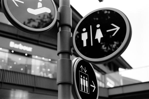 Fujifilm NEOPAN F (FF)

##English
A black-and-white image of three circular directional signs mounted on a pole. The top sign features a symbol of a person standing on an escalator. The middle sign shows a man and woman icon with an arrow pointing right, indicating restrooms. The bottom sign depicts an elevator symbol with an arrow pointing right. In the background, there is a building with the word "Barbour" visible.
##中文
一张黑白照片，显示三个圆形方向标志固定在一根杆子上。最上面的标志是一个站在手扶电梯的人的图标。中间的标志是男女图标，右侧有一个箭头，表示洗手间。…