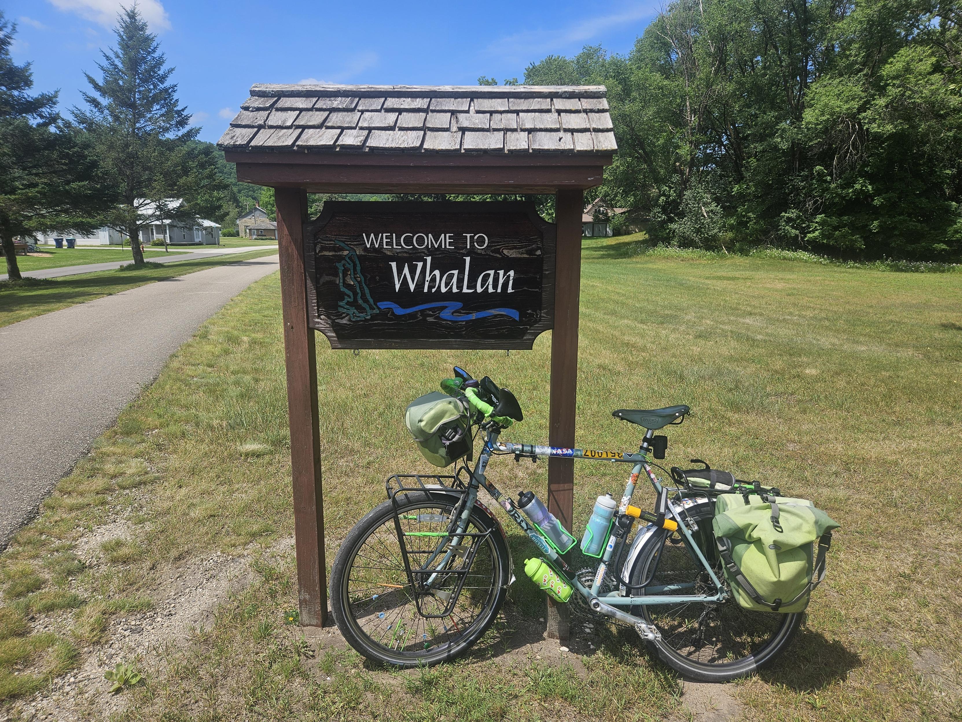 A bicycle with green bags leans against a sign that says Welcome to Whalan, bext to an asphalt trail. Green grass freshly mowed on the ground. Green trees an a blue sky in the background.