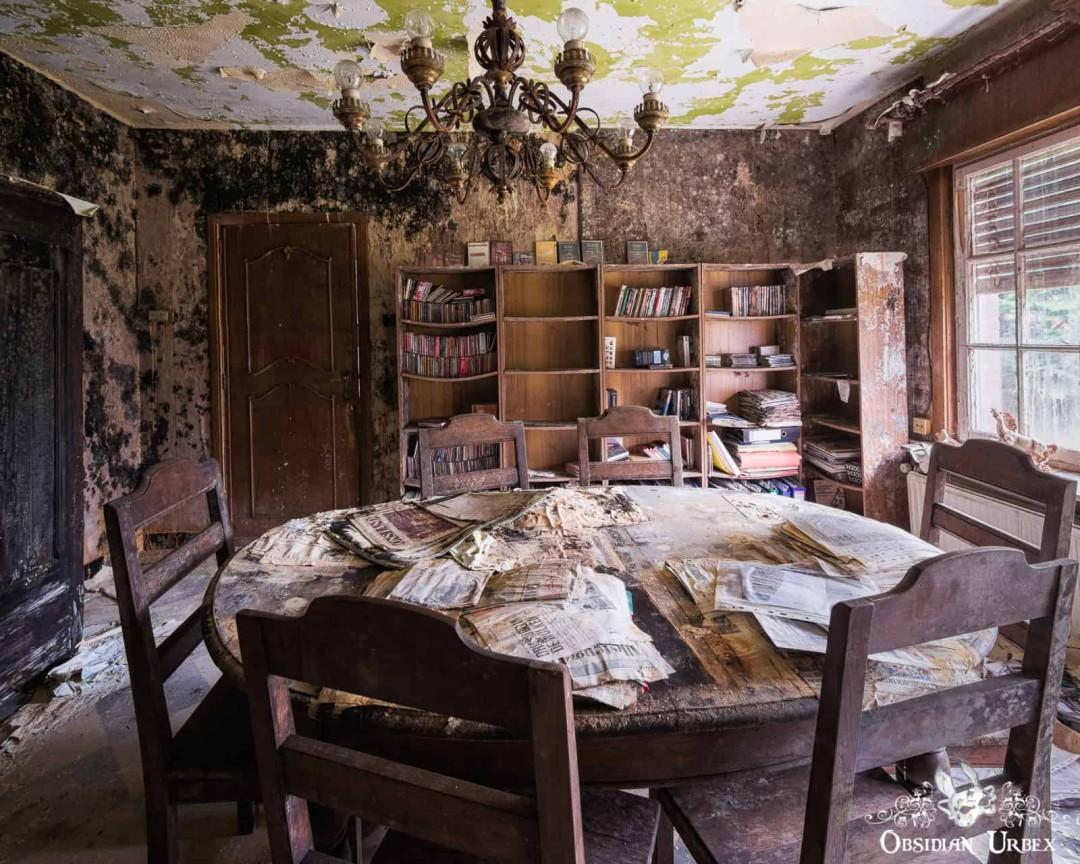 An abandoned, mold-covered room features a round table with decaying papers. Bookshelves hold books and records.