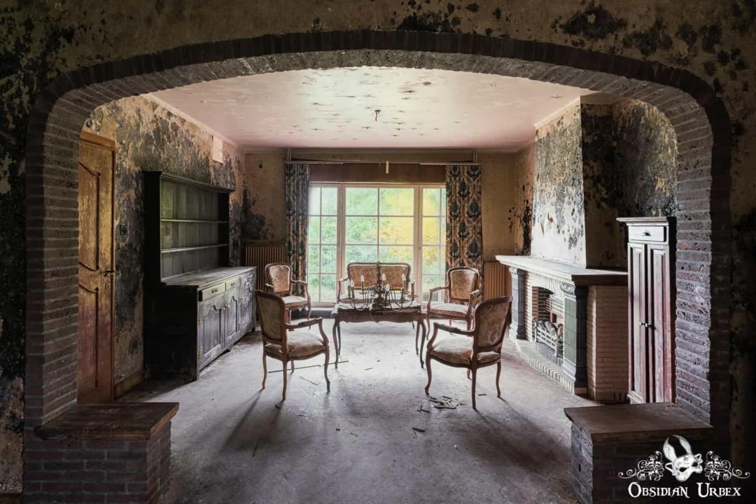 A brick archway frames a decaying living room with period furniture, peeling walls, and sunlight streaming from a large window.