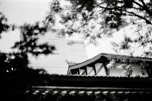 Fujifilm NEOPAN F (FF)

English
A black-and-white photograph capturing a partial view of a modern building with a grid-like facade, partially obscured by tree branches and a traditional east Asian building in the foreground. The building's upper section is visible, with a triangular architectural element protruding from its side. The sky is bright, creating a high-contrast silhouette effect.
中文
一张黑白照片，捕捉到一栋现代建筑的部分视图，其立面呈网格状，前景被树枝和一栋传统东亚建筑部分遮挡。建筑的上部可见，侧面有一个三角形的建筑元素突出。天空明亮，形成高对比度的剪影效果。