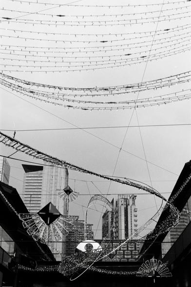 Fujifilm NEOPAN F (FF)

English
A black-and-white photograph of a cityscape featuring tall buildings and a network of overhead cables. Decorative elements, including diamond-shaped and fan-shaped light fixtures, hang from the cables. The image captures an urban scene with a focus on architectural and decorative details.
中文
一张黑白城市风景照，展现了高楼大厦和密集的架空电缆。装饰性元素，包括菱形和扇形的灯具，悬挂在电缆上。这张图片捕捉了城市景观，重点展现了建筑和装饰细节。