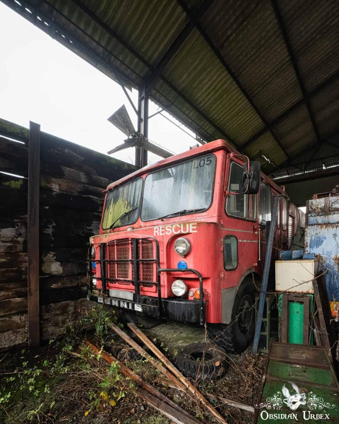 An abandoned red airport crash tender fire truck, marked "RESCUE", sits in a derelict shed. Overgrown plants surround the vehicle, indicating its long disuse.