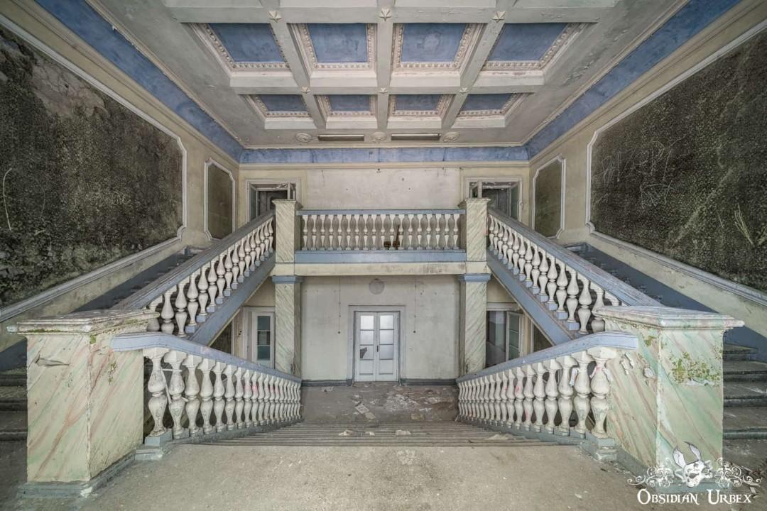 A grand, decaying double staircase ascends symmetrically in an abandoned building with a coffered blue ceiling.
