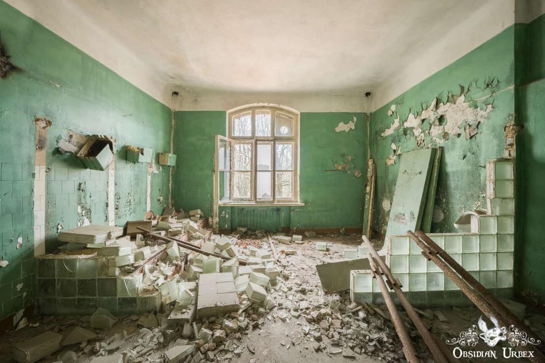 A derelict room with peeling green walls and debris from broken tiles. A central window brightens the abandoned space.