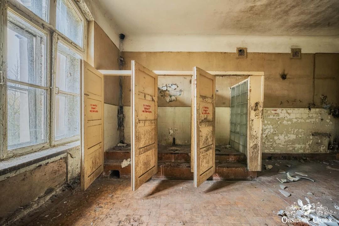 An abandoned washroom featuring three open stalls with decaying walls, dirty floors, and large windows.