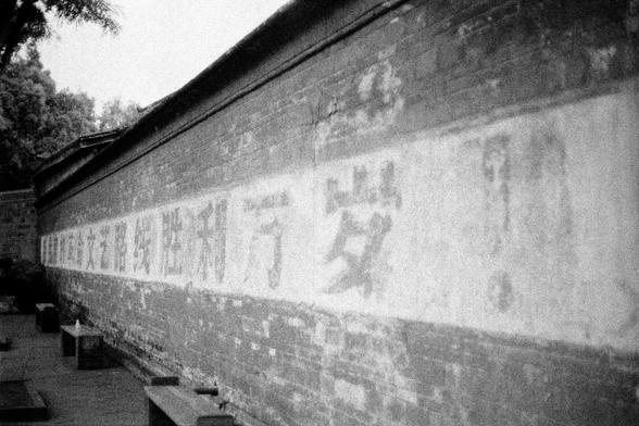 Lucky LUCKY SHD 400 (FF)

English
Black-and-white photograph of a long, curved brick wall with faded Chinese characters inscribed on its surface. The wall stretches horizontally across the image, with trees visible in the background. The characters are large and appear weathered, blending into the texture of the bricks. The scene is quiet, with a few stone benches placed along the base of the wall.
中文
黑白照片，展现了一面长长的、弯曲的砖墙，墙面上刻有褪色的中文字。砖墙横贯整个画面，背景中可见树木。这些字体较大，看起来经过了风化，与砖块的纹理融为一体。场景安静，墙脚下放置着几张石凳。