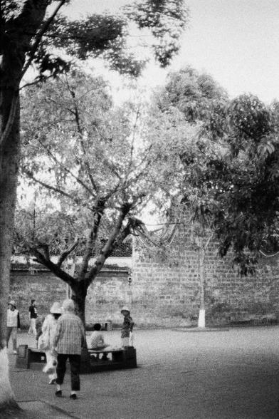 Lucky LUCKY SHD 400 (FF)

English
Black-and-white photograph of a park scene. A large, leafless tree dominates the left foreground, its branches spreading outward. In the background, a tall brick wall runs horizontally. Several people, some wearing hats, are scattered across the open space. Two individuals stand near a bench, while others walk or stand in small groups. The ground is paved, and the scene is bathed in natural light, casting soft shadows.
中文
黑白照片，展现了一个公园场景。左前方有一棵大树，枝干茂密但没有叶子。背景是一面…