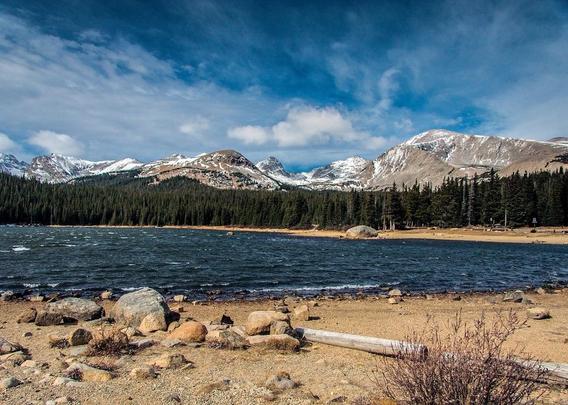 Indian Peaks Range behind Brainard Lake. Along the Front Range of the Rocky Mountains in Colorado. A rock-strewn sandy beach in front of a deep-blue lake with white cap waves in a stiff breeze. The far shore is backed by (probably) Ponderosa pine trees, behind which is a row of snowy mountain tops. Above all is a dark blue sky with white clouds. August 2013.