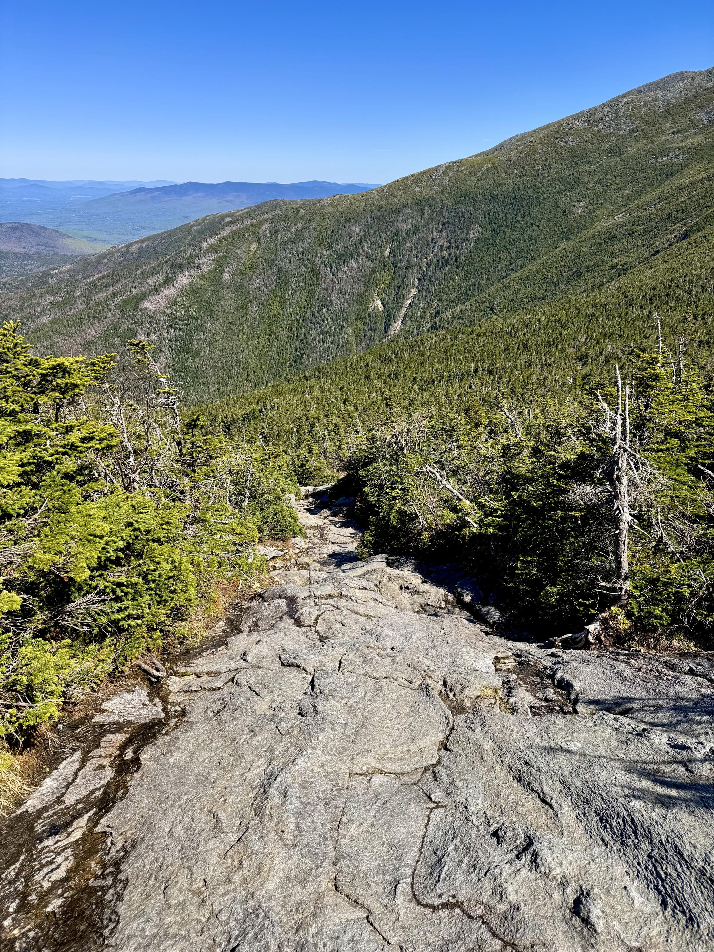 Photo of steep rock slab as viewed from top looking down.  The slab is the "trail" that leads up the mountain ravine.  Mountain ridges and peaks seen off in the distance.  Blue sky overhead.

This is Ammonoosuc Trail as I am about to get above the treeline.  I took this photo on May 22, 2026 during my hike in the Presidential Range, White Mountains, NH.