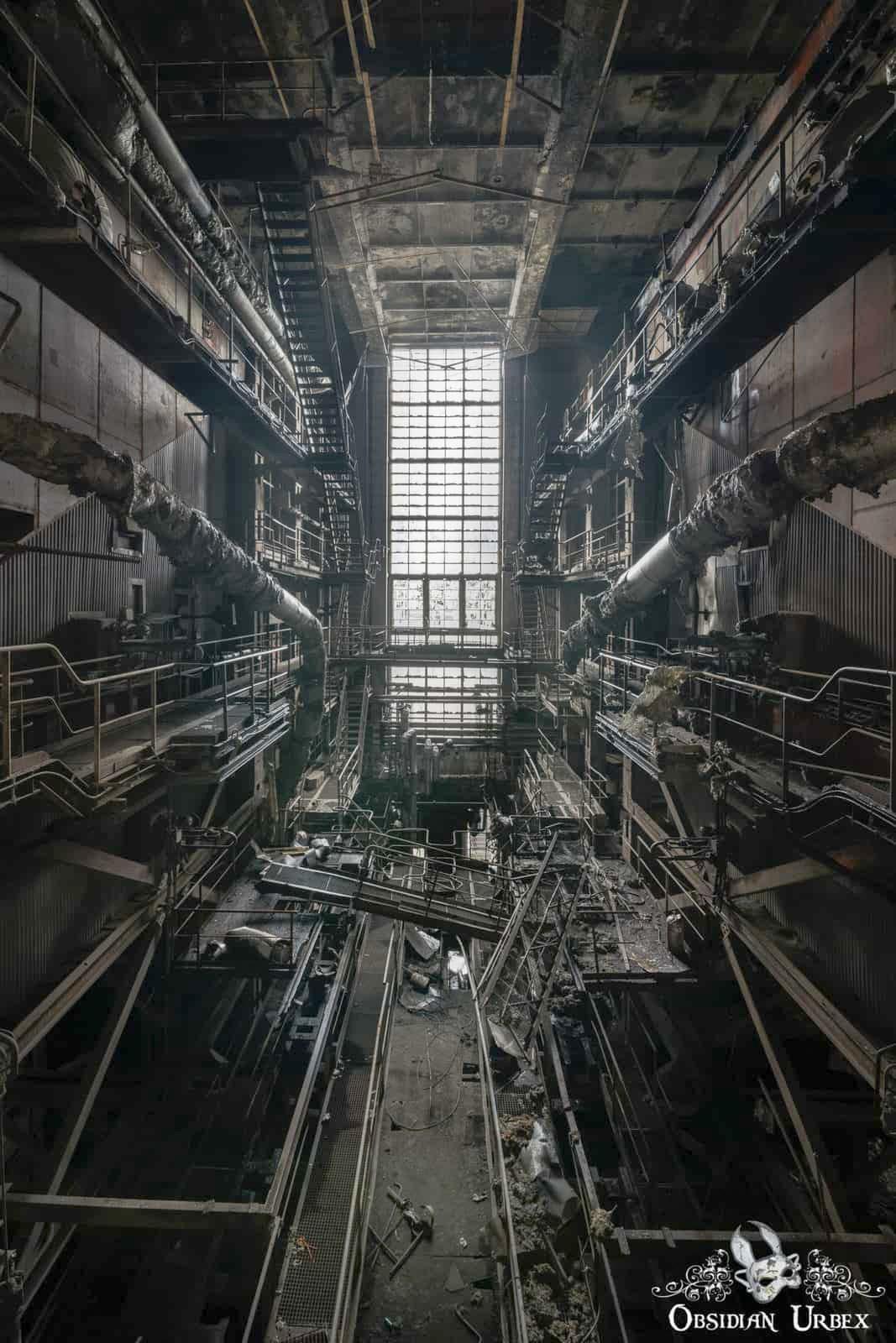 A vast, decaying industrial area with multiple levels of pipes, catwalks, and a bright window,