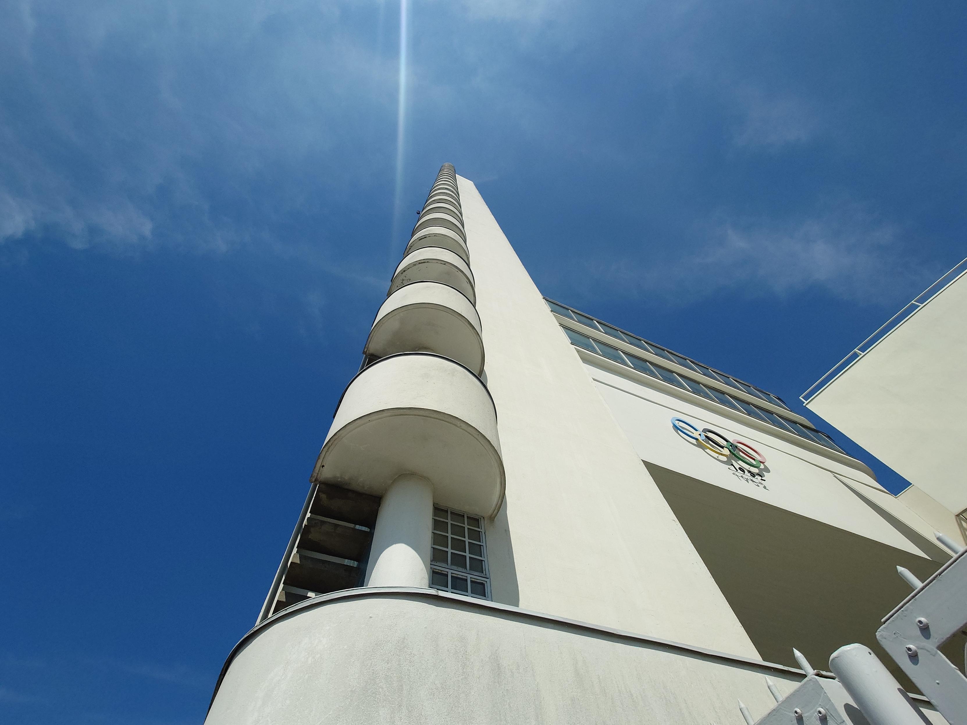 A view up the tower from below shows semicircular architectural elements structuring a staircase up the tower, with the Olympic rings on an adjoining wall just too the right. It is all in white, slightly off-colour, against a blue sky with just whisps of cloud.