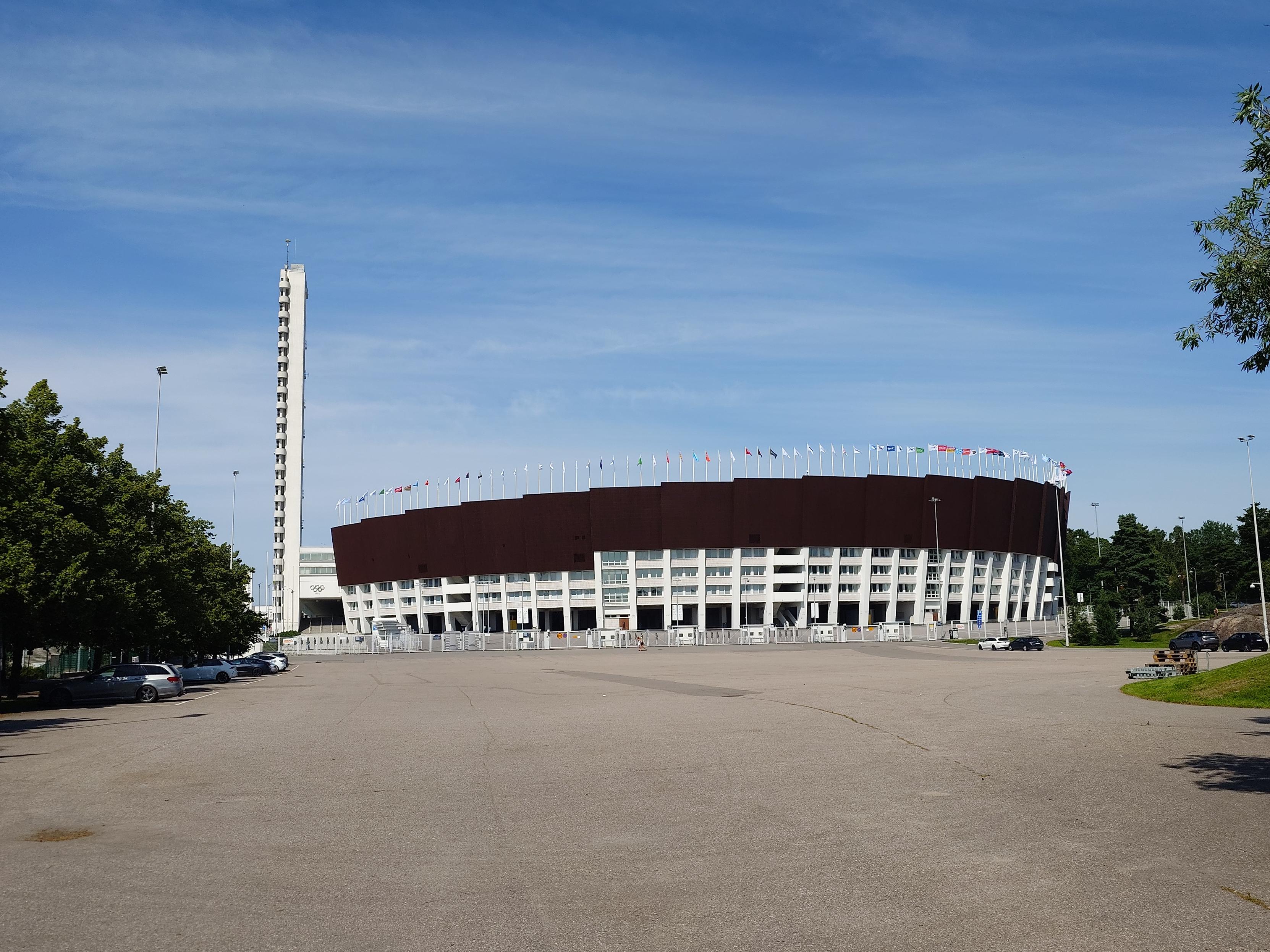 An overall view of the stadium shows it's basket-like white understructure topped by brown panelling to form a large oval behind an empty square flanked by trees, and sporting the iconic stadium tower, incredibly slender and very high, and all white.