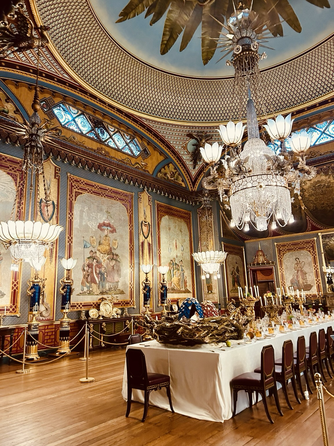A table laid out for a royal feast in the banquet in hall. A huge chandelier above the table wrights a tonne. Multiple additional hanging lights. Opulent oriental style decoration on walls.