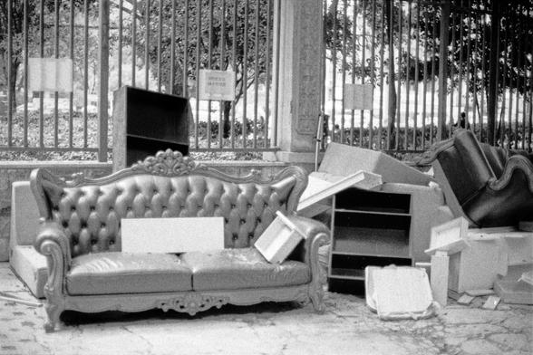 Lucky LUCKY SHD 400 (FF)

English
A black-and-white photograph showing an outdoor scene with discarded furniture. In the foreground, there is an ornate, tufted sofa with curved armrests and wooden legs, placed on a paved surface. Surrounding the sofa are various items: a wooden cabinet, a shelving unit, a rolled-up mattress, and several cardboard boxes, some of which are open. In the background, there is a metal fence with vertical bars and trees visible beyond it. A sign with the text is parti…