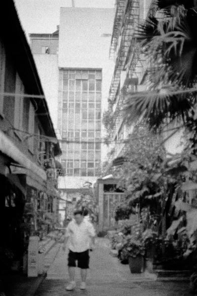 Lucky LUCKY SHD 400 (FF)

English
A black-and-white photograph of a narrow alleyway flanked by buildings. A person in a white shirt and dark shorts is facing the camera. The alley is lined with potted plants and greenery, and a tall building with many windows is visible in the background.
中文
一张黑白照片，展示了一条狭窄的小巷，两侧是建筑。一个穿着白色上衣和深色短裤的人正面对着镜头行走。小巷两侧种满了盆栽植物和绿化带，背景中可见一栋高层建筑，有许多窗户。