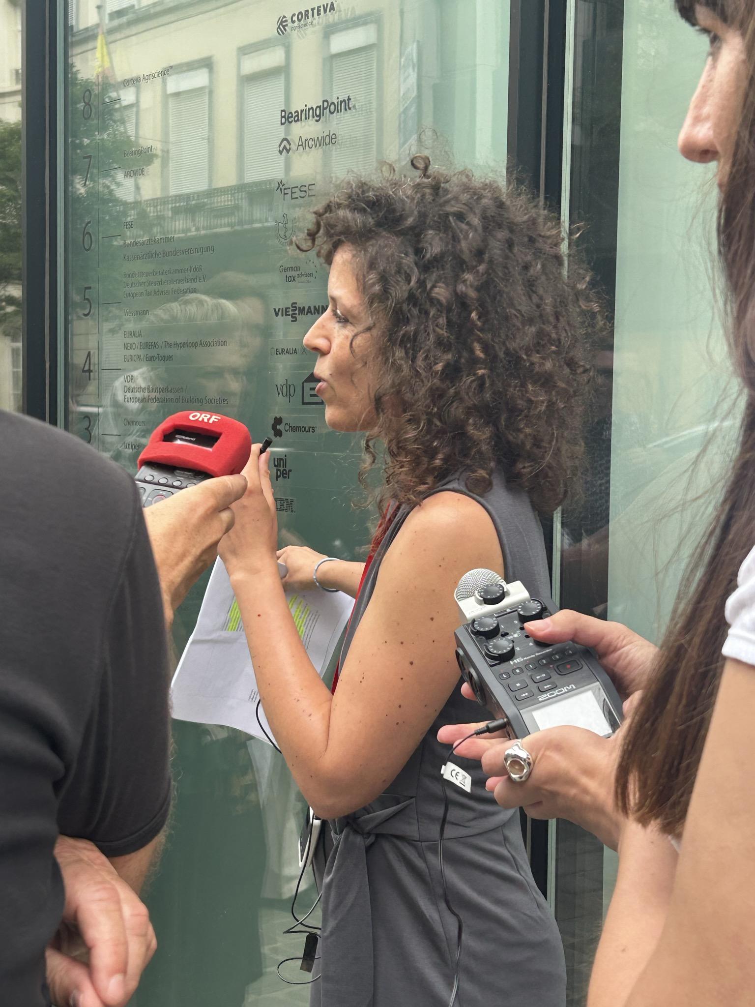 A woman with curly dark hair speaks into an ORF microphone while holding papers, standing in front of a glass building directory listing companies including Corteva Agriscience, BearingPoint, Chemours, and Uniper. A person beside her holds a Zoom audio recorder. The scene appears to be an outdoor press interview in Brussels.