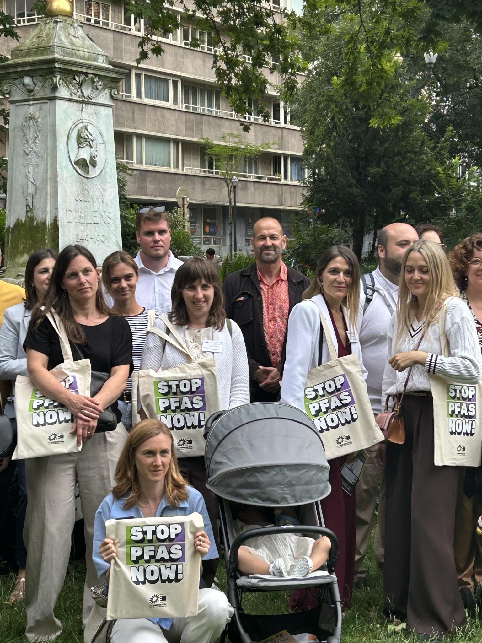 A closer shot of a group of around 12 people posing in a park in front of a stone monument. Several are holding tote bags printed with 'Stop PFAS Now!' One person is seated in the foreground holding a bag, with a pram next to them. The monument behind the group bears a carved relief portrait.