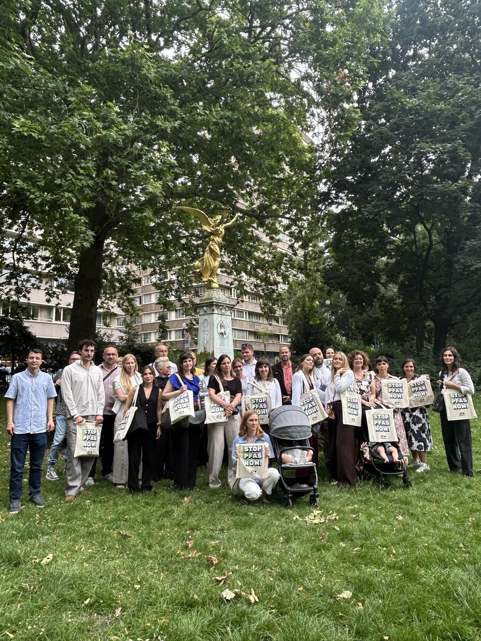 A large group of around 25 people pose together outdoors in a park, standing in front of a stone monument topped with a golden winged statue. Most participants are holding tote bags printed with 'Stop PFAS Now!' One person is seated on the grass holding a bag, with a pram beside them.