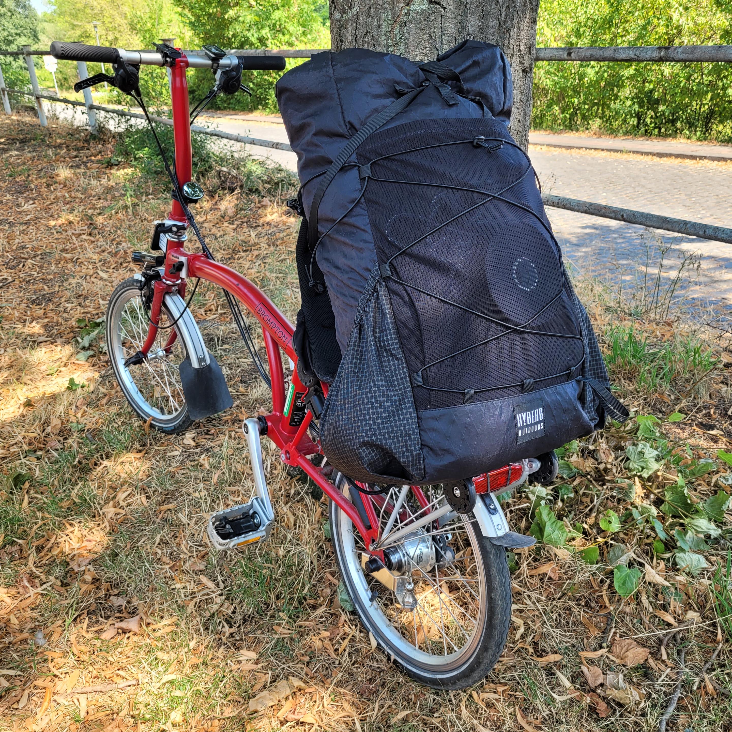 Foto eines roten Brompton Faltrads, das an einem Baum lehnt. Auf dem hinteren Gepäckträger steht ein großer Rucksack schräg nach vorne geneigt.