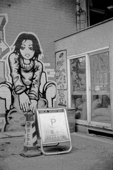 Lucky LUCKY SHD 400 (FF)

English
A black-and-white photograph of a street scene featuring a large mural of a cartoonish female character sitting on the ground, leaning forward with her hands on her knees. The mural is on the side of a building. In the foreground, there is a metal A-frame sign with the letter "P" and Chinese characters. The background includes a storefront with glass doors and additional graffiti.
中文
一张黑白色调的街道场景照片，照片中有一幅大型壁画，画的是一个卡通风格的女性角色坐在地上，双手放在膝盖上，身体前倾。壁画位于建筑的侧面。前景中有一个金属A型标…