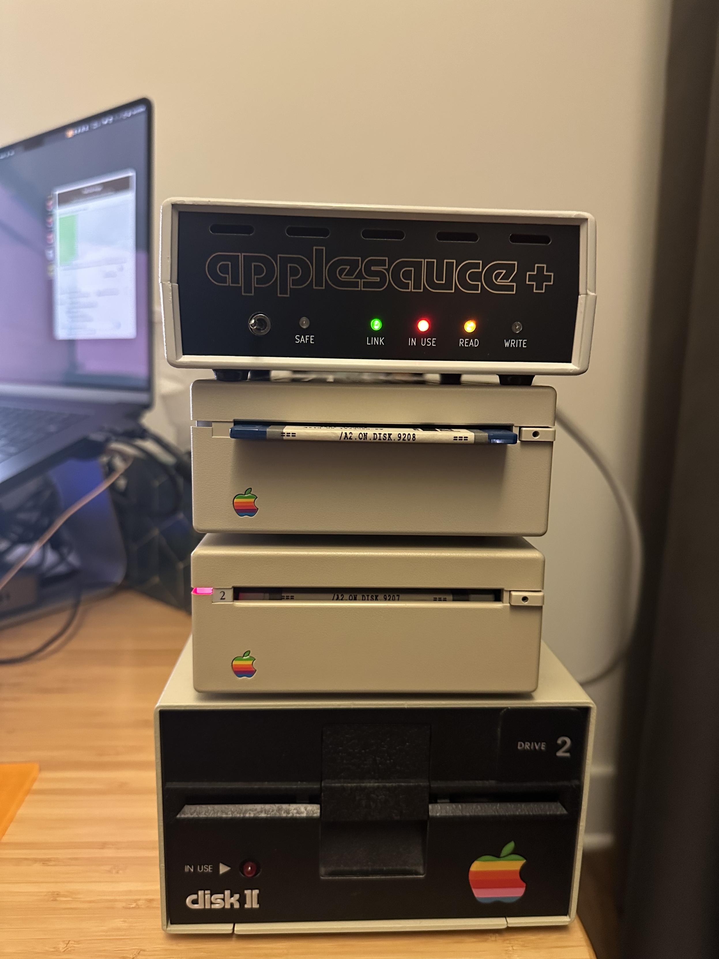 Applesauce+ at the top of a stack containing 2 3.5 inch Apple external floppy drives and an Apple Disk II. Visible in the background is a MacBook Pro running the Applesauce software and miscellaneous cables.
