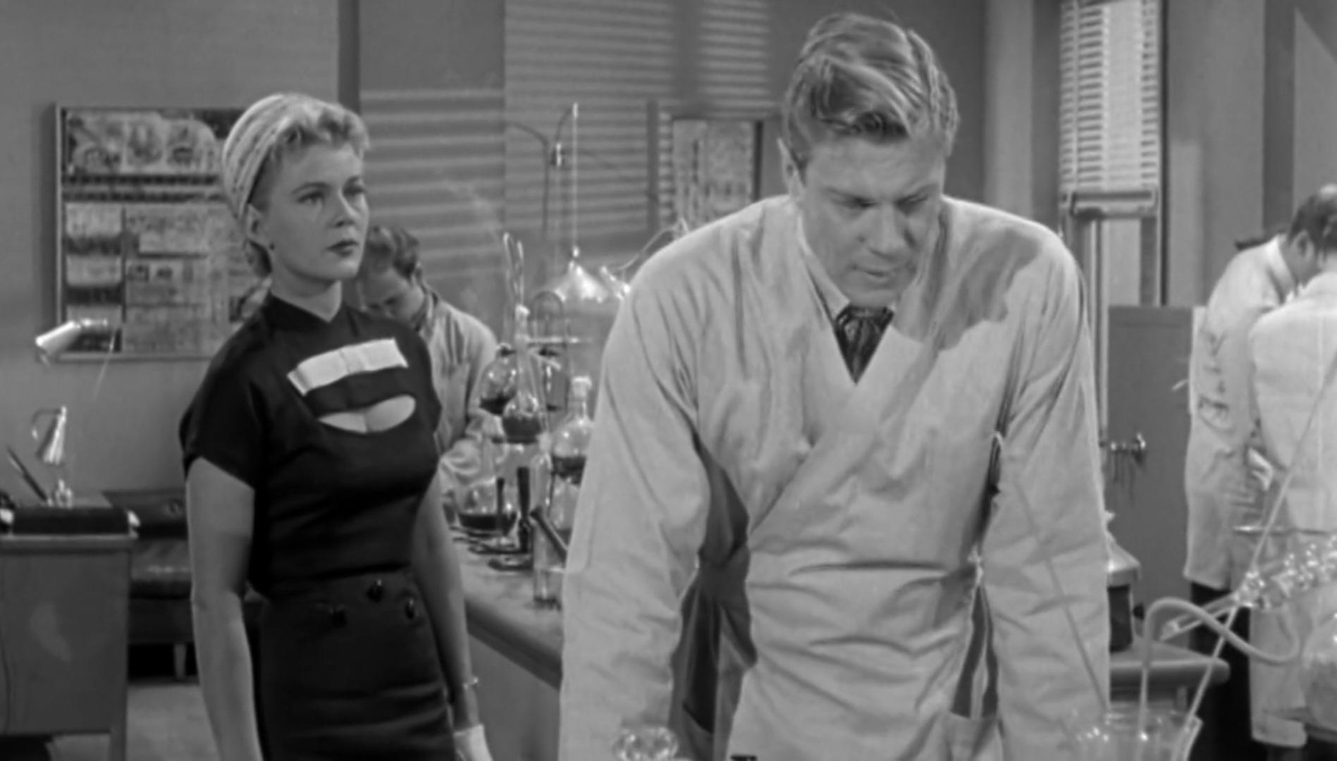 Actress Peggie Castle, wearing a black blouse showing cleavage, in the middle of a science lab. Peter Graves looks away, trying not the stare.