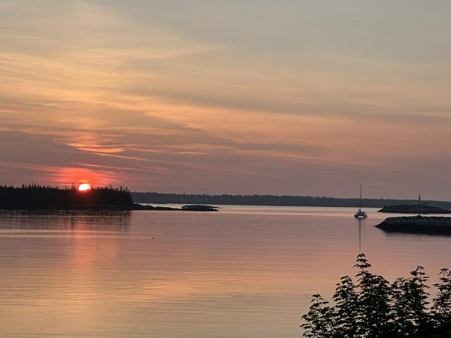 Sun rising over ocean water, in Nova Scotia. Sail boat and a break water on the right. Sun rising over tree line , on the left