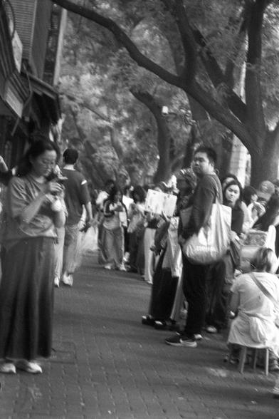 Ilford FP4 Plus 125 (FF)

English
A black-and-white photograph of a busy outdoor scene with a line of people standing under large trees. The individuals appear to be waiting or queuing, some holding bags or reading. The trees provide a canopy, and the background includes a building with a striped awning. The atmosphere suggests a public gathering or event.
中文
一张黑白照片，展示了一个繁忙的户外场景，一群人在大树下排队等待。有些人拿着袋子或在阅读。大树形成天然的遮蔽，背景中有一栋带条纹遮阳棚的建筑。氛围暗示这是一次公共集会或活动。