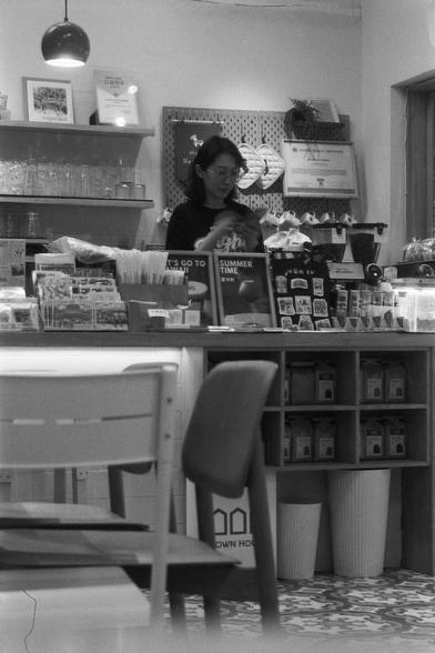 Ilford FP4 Plus 125 (FF)

English
A black-and-white interior shot of a shop or café. A person stands behind a counter filled with various products, including boxes, bags, and displayed items. Shelves in the background hold more merchandise, and a sign reading "SUMMER TIME" is visible. The space is well-lit, with a hanging lamp and a chair in the foreground.
中文
一张黑白室内照片，拍摄的是一家商店或咖啡馆。一人站在柜台后，柜台上摆放着各种商品，包括盒子、袋子和陈列品。背景中的架子上也放着更多商品，还有一个写着“SUMMER TIME”的招牌。空间光线充足，前景有一盏吊灯和一把椅子。