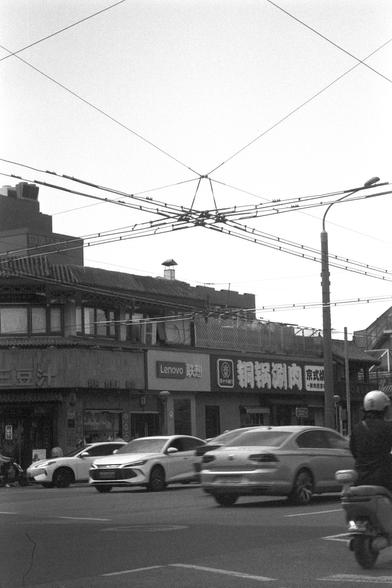 Ilford FP4 Plus 125 (FF)

English
A black-and-white street scene featuring a two-story building with traditional Asian architectural elements. The facade includes a tiled roof, wooden windows, and signage in Chinese characters. Overhead, a dense network of electrical wires crisscrosses the sky. In front of the building, several cars are parked or driving on the street, and a motorcyclist with a helmet is visible on the right side.
中文
一张黑白街道照片，呈现一栋两层楼的传统亚洲风格建筑。建筑立面有瓦片屋顶、木制窗户，以及中文字样的招牌。上方是密集交叉的电线…