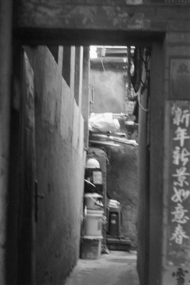 Ilford FP4 Plus 125 (FF)

English
A black-and-white photograph showing a narrow alleyway framed by stone walls. The right wall features vertical Chinese characters. The alley leads to a dimly lit interior space with stacked objects, including barrels and bags. The scene captures a sense of depth and texture, with the rough stone walls and the play of light and shadow.
中文
一张黑白照片，展示了一条由石墙围成的狭窄小巷。右侧的墙上有竖排的中文字。小巷通向一个光线昏暗的内部空间，里面堆放着桶和袋子等物品。画面展现了深度和质感，粗糙的石墙与光影的交织营造出特有的氛围。