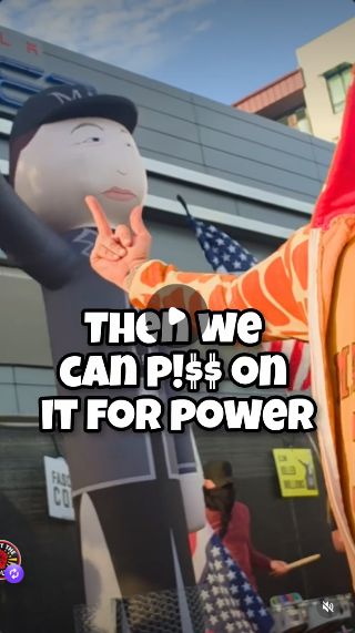 Still of the video from comedian Robby Roadsteamer, who is flipping off the diner in his Jeffrey Epstein Giraffe costume. Outside the Tesla Diner you see a scaffold with black fabric hanging over it so that diners don't see the protesters, although they sure as hell must hear them. There is a guy with a full drum set playing drums next to Inflatable Saluting Elon, which is like those dancing inflatable figures outside car dealerships except it throws Nazi salutes. There are US flags and signs that protestors have hung on the wall.