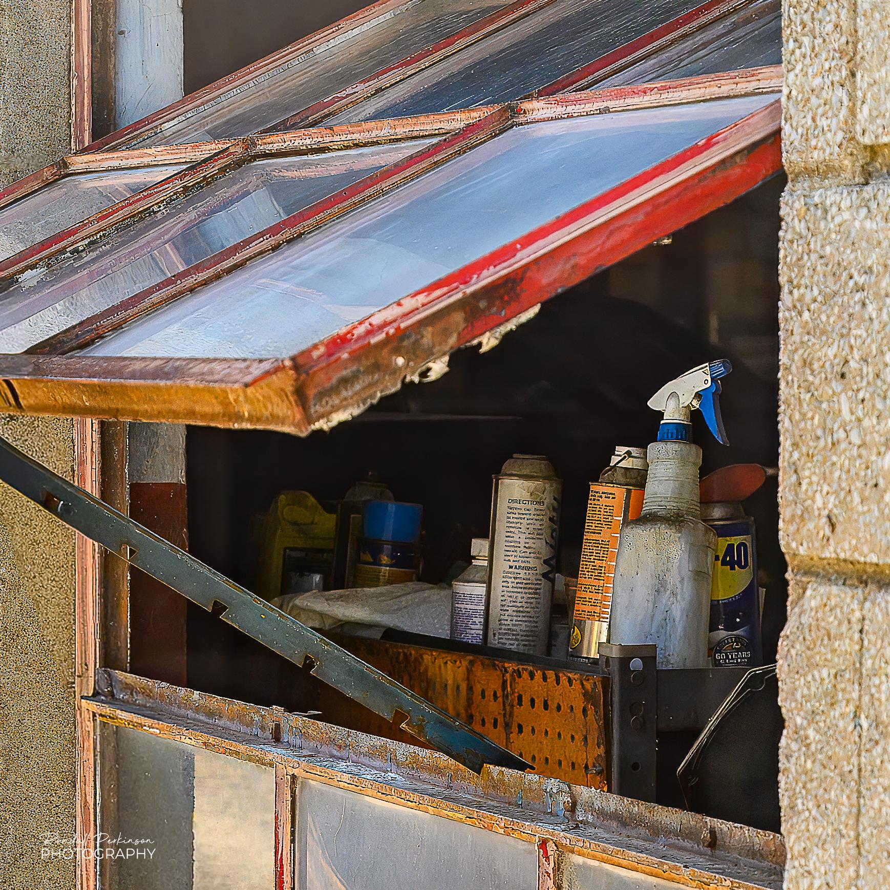 An auto repair shop window is rotated open and propped up with a metal bar.  Inside you can see a spray bottle and cans similar to spray paint cans.