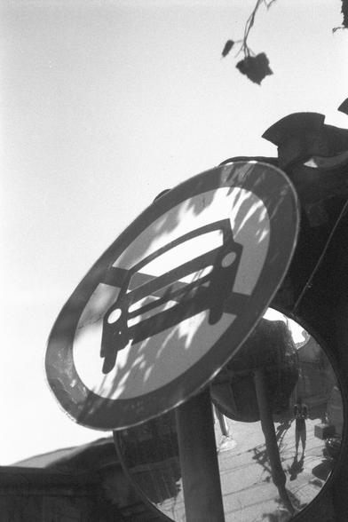 Ilford FP4 Plus 125 (FF)

English
A black-and-white close-up photograph of a circular traffic sign and a reflection mirror behind it. The sign features a symbol of a car and a line cross it diagonally, indicating cars are forbidden. The sign is slightly tilted and reflects light, creating a glare effect. The mirror reflects  the alleyway and the photographer.The background is blurred, focusing attention on the sign.
中文
一张黑白特写照片，展示了一个圆形交通标志和它背后的反光镜。标志上有一辆车和一个向下的斜线，表示车辆禁止通行。标志略微倾斜，并反射光线，产生了眩光效果。反…